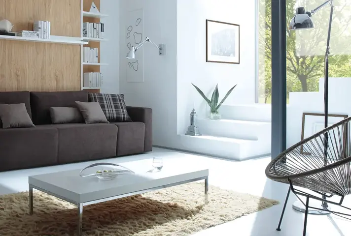 Helles Wohnzimmer mit braunem Sofa vor Holzrückwand und Wandregalen, rechts weisse Stufen mit Pflanze, davor weisser rechteckiger Couchtisch auf beigem Hochflorteppich.